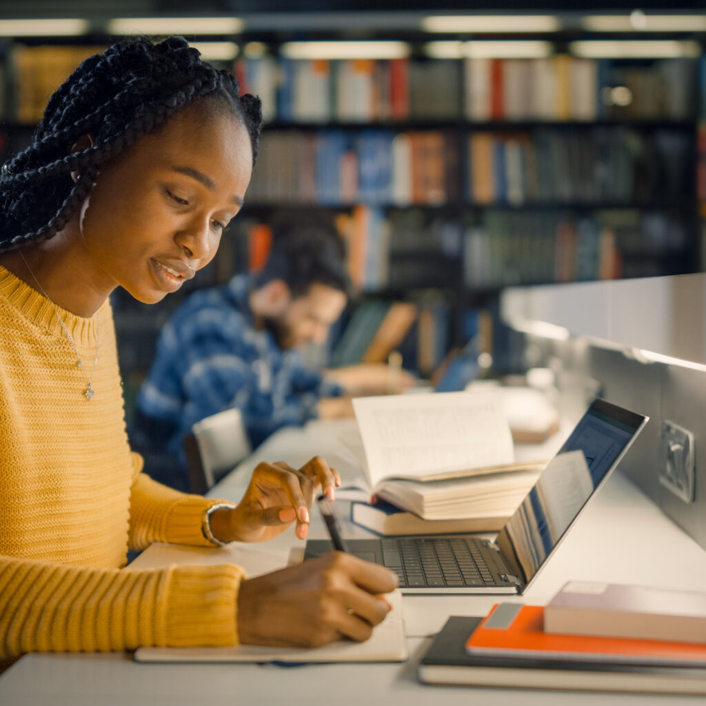 University Library: Gifted Beautiful Black Girl Sitting at the Desk, Uses Laptop, Writes Notes for the Paper, Essay, Study for Class Assignment. Diverse Group of Students Learning, Studying for Exams.